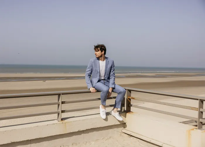 un homme en costume bleu ciel, il porte des sneakers et un t-shirt blanc, il est sur une plage au bord de la mer